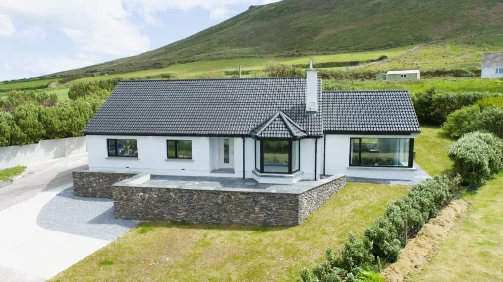 White bungalow with a dark tiled roof and stone wall, set against a green hillside